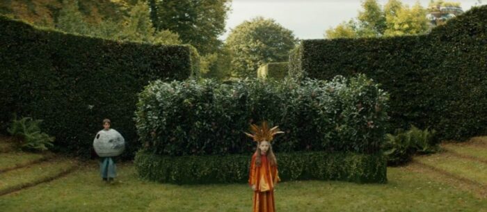 A little girl with a corwn on stands in front of trimmed hedges