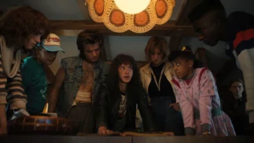 Nancy, Dustin, Steve, Eddie, Robin, Erica, and Lucas look at a book on a table while Max stands to the side