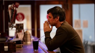 Nathan Fielder sits at a bar with his hands together to the front of his face