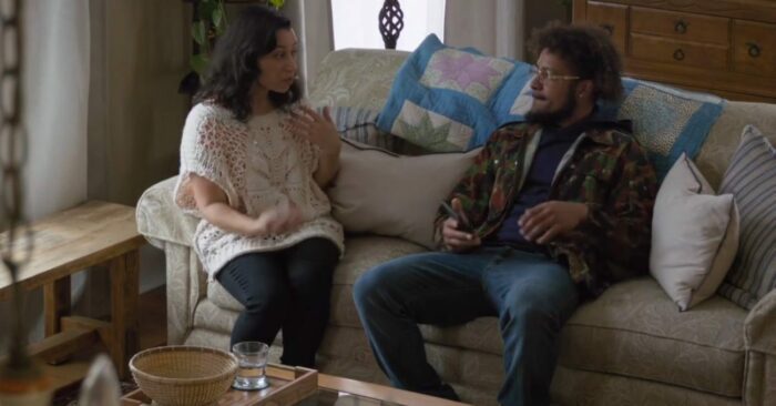 Angela and Robin sit on a couch. She gestures with her hands while talking to him