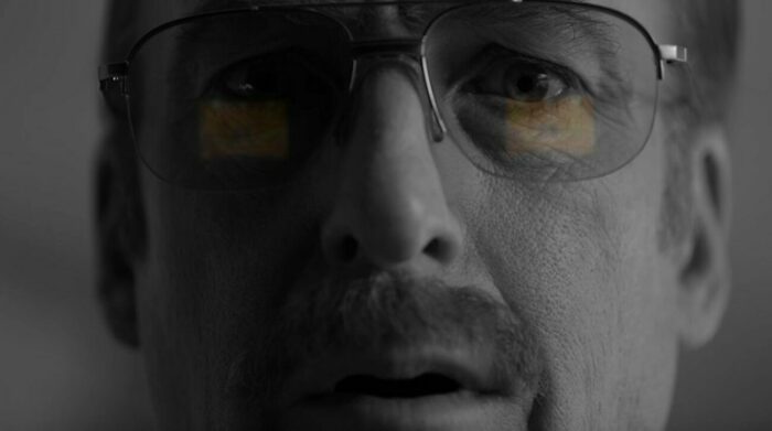 A black and white closeup of Gene with the reflection of Saul Goodman TV commercial in his glasses in color