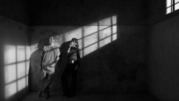 A black and white image of Jimmy and Kim leaning against the wall in prison with the shadows from the window bars on the wall