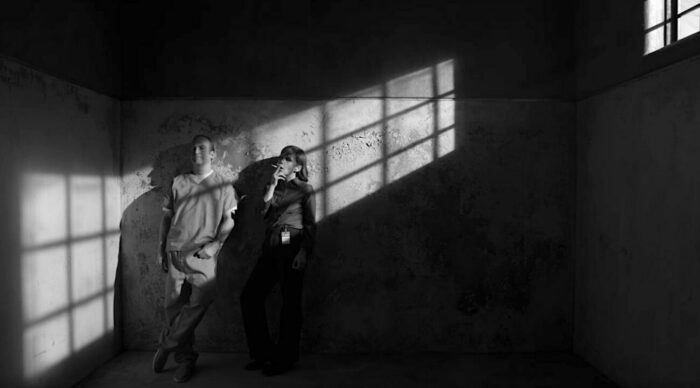 A black and white image of Jimmy and Kim leaning against the wall in prison with the shadows from the window bars on the wall