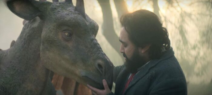 A bearded man in a suit gently cradles the head of a dragon-like creature in his hands.