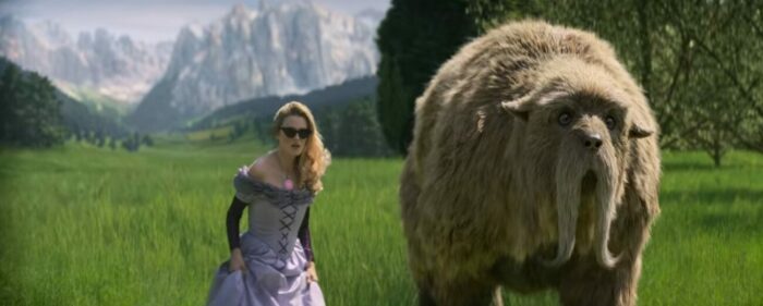 A woman with long blonde hair wearing dark sunglasses and a purple gown stands in a grassy field with a large furry bear-like creature.