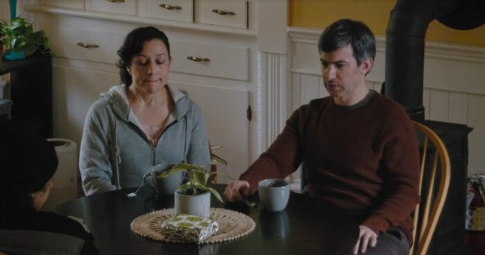 Angela and Nathan sit at a kitchen table, looking glum