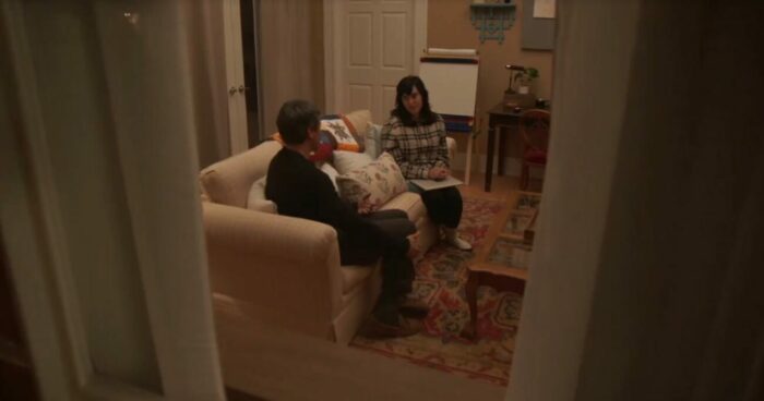 Nathan and Fake Angela sit on a couch, in a shot through a doorframe