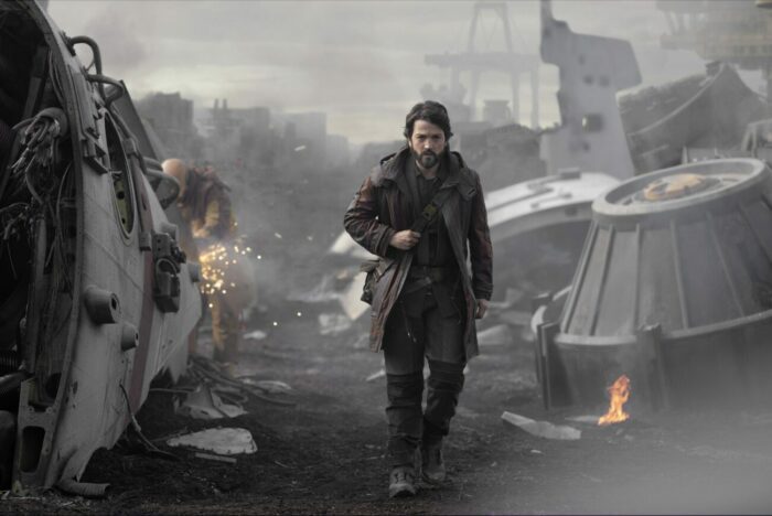 Andor walks through a yard surrounded by wreckage and such