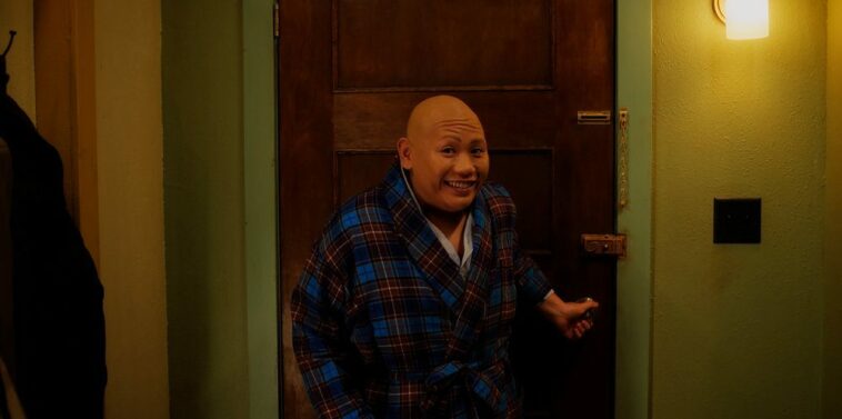Jacob Batalon as Reginald smiling to the camera in his rob at the door to his apartment after seeing that Sarah is just outside