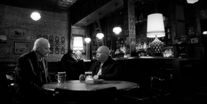 Bernard Cuffling as Jenkins, Jacob Batalon as Reginald in the black and white dive bar sitting at a table having a drink