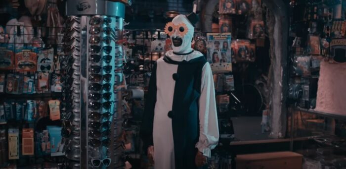 Art the Clown wears a pair of silly sunglasses that look like gears, while standing beside a rack of sunglasses, in Terrifier 2