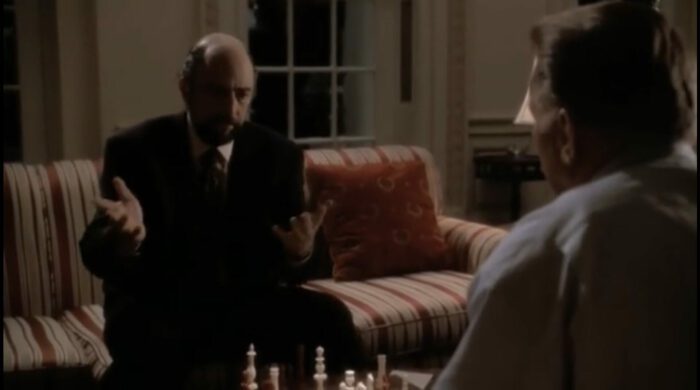 Toby facing the camera gesticulating as President Bartlet watches him across the chess board