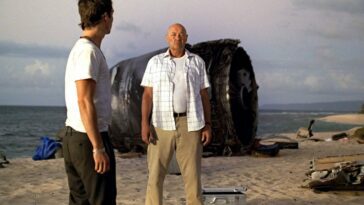 Locke (Terry O'Quinn) standing in front of part of the crashed plane on a beach with Boone to the left
