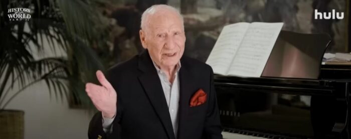 Mel Brooks wears a dark suit and extends a hand to introduce History of the World Part II in the trailer for the show