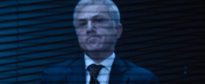 Regus makes a face with downturned lips as he seems to look at a camera through blinds in The Consultant