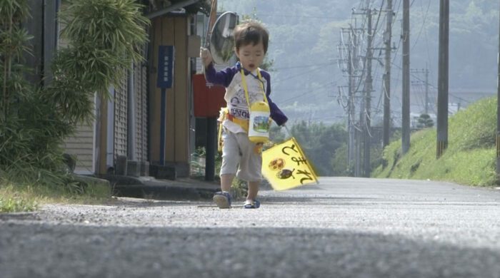A Japanese boy carrying flags and other items makes the long walk home, toward camera.