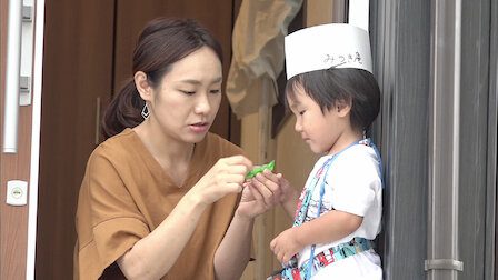 A woman prepares her young son, who is wearing a sushi chef's hat, for his first errand.