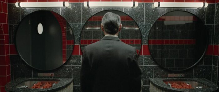 Jack, his back to the camera, in the bathroom featuring three circular mirrors and sinks with red spongey hand cleaners in them in Hello Tomorrow! S1E6 "The Numbers Behind the Numbers"