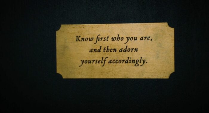 A placard reads, "know first who you are, and then adorn yourself accordingly"