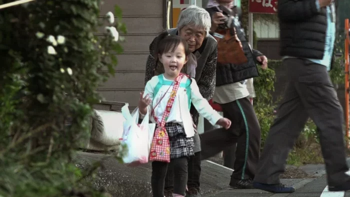 A young Japanese girl joyfully departs from the elderly seamstress and shopkeeper who helped her.