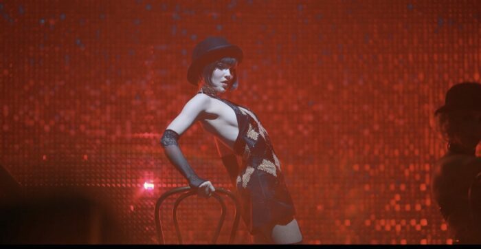 Jenny dancing in front of a red curtain, wearing a black hat and holding onto a chair