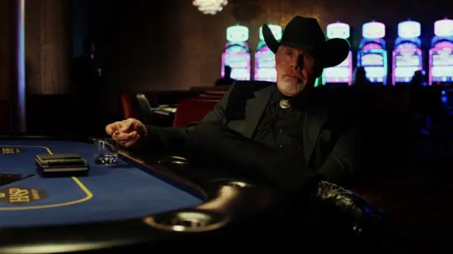 Frost Sr. sitting at a card table wearing a black suit and hat, holding a glass of whiskey