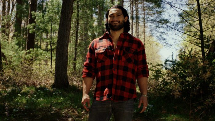 A brawny mountain man in a red and black plaid shirt standing in the woods in the golden sunlight