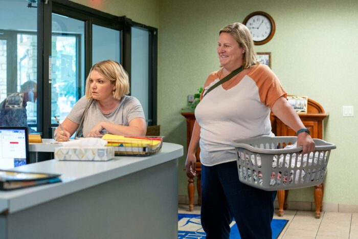 Sam and Tricia stand at a receptionist's desk. Sam is holding a laundry basket