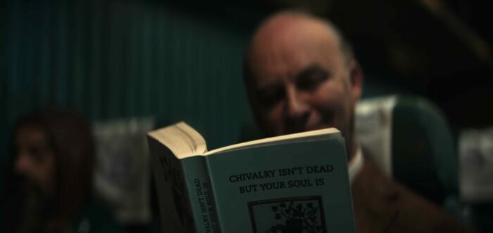 Mrs. Davis S1E3 - Apron Man sits in a train car reading a book titled "Chivalry Isn't Dead but Your Soul Is"