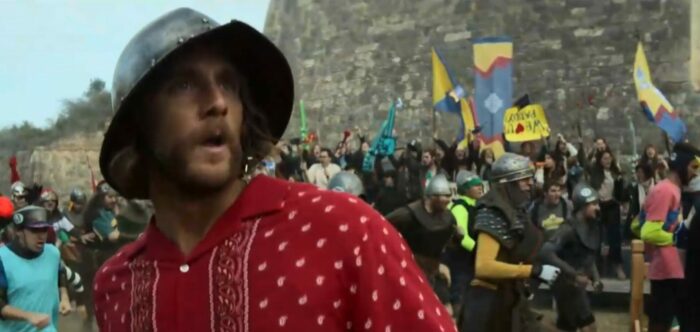 Wiley in a medieval helmet stands aghast while the crowd of participants races behind him