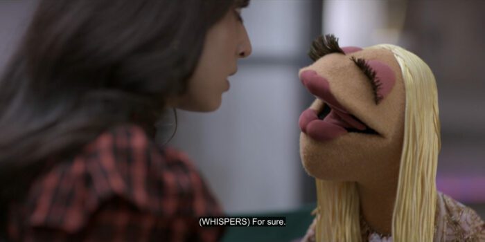 Janice whispers, "Fer sure," to Hannah Singh (Saara Chaudry), in the TV show, "The Muppets Mayhem."