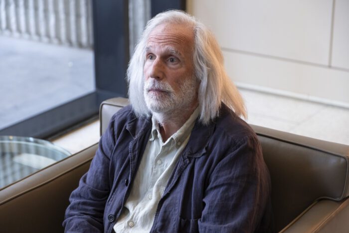 Gene sits in a chair with long white hair and a beard