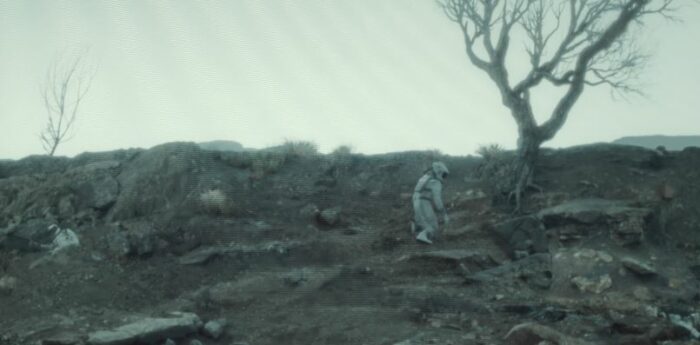 A figure in a spacesuit stumbles near a tree