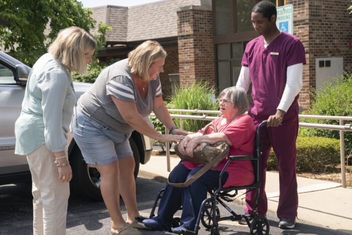 Sam and Tricia hunch over to say hello to MJ as she is pushed out to meet them in the parking lot in a wheelchair