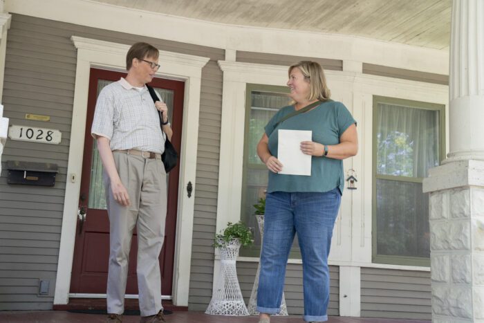 Sam and Joel on the porch of a house. She holds a piece of paper and he has a bag over his shoulder