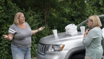Sam gestures with her arms wide to Tricia, who stands on the other side of a car's hood that has cups and a bag resting on it in Somebody Somewhere S2E4