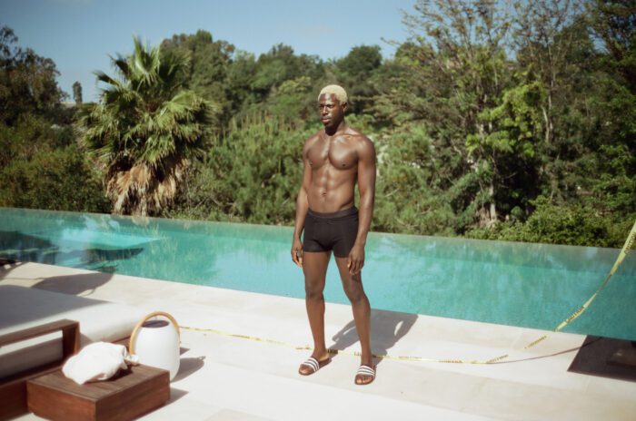 Moses Sumney plays Izaak in The Idol. In this shot, he stands beside an infinity pool in trunks and sandals. 