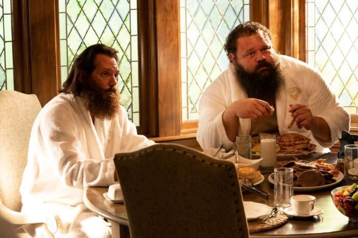 Chuck and Carl, wearing bathrobes, sit at a table eating breakfast. 