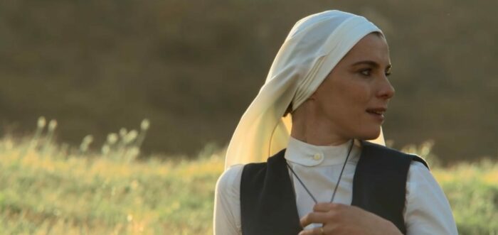 Mrs. Davis S1E4 - Sister Simone stands in a field smiling and holding her cross necklace in her hand