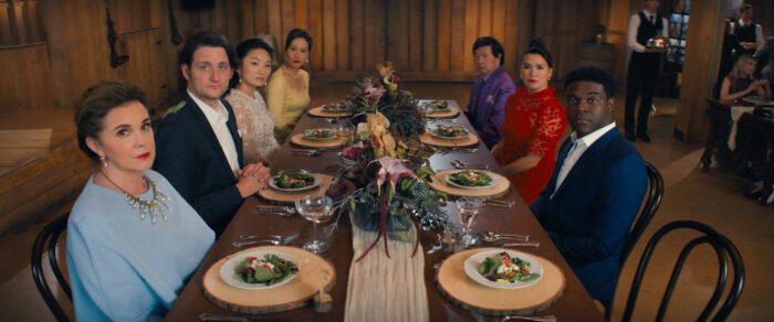 The Afterparty S2E4: L-R Isabel (Elizabeth Perkins), Edgar (Zach Woods), Grace (Poppy Liu), Vivian (Vivian Wu), Feng (Ken Jeong), Zoe (Zoe Chao) and Aniq (Sam Richardson) sitting around a dinner table looking directly to the camera, i.e. Hannah's perspective. 