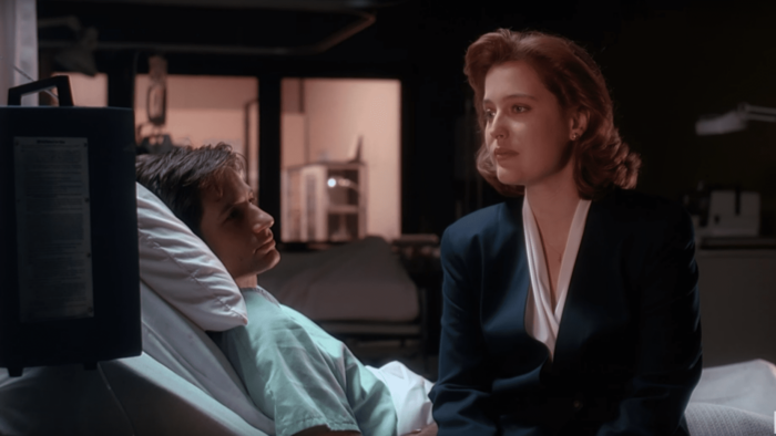 Fox Mulder lays in a hospital bed wearing a faded teal hospital gown. Dana Scully, wearing black blazer, sits on the edge of the bed and looks off to the left. She appears contemplative, and Mulder listens intently.