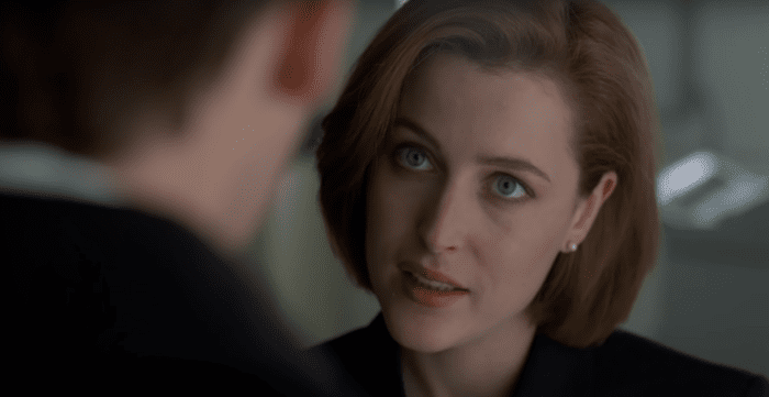 Special Agent Dana Scully, a woman with short red hair, looks up into the face of a man whose profile is blurred. She wears an inquisitive look on her face, as if she is asking an important question.