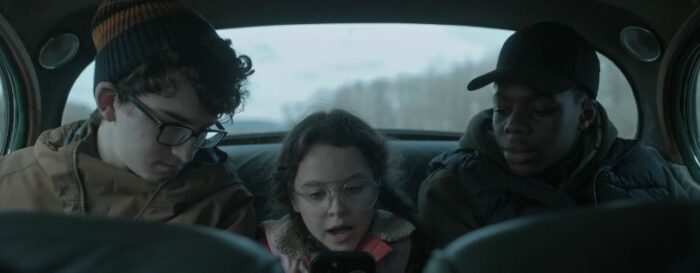 Penny sits in the backseat of a car looking at a smartphone, with Alfie and Darwin to each side of her