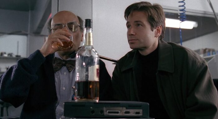 Mulder and Dr. Ivanov sit with a bottle of whiskey between them, as Ivanov takes a drink