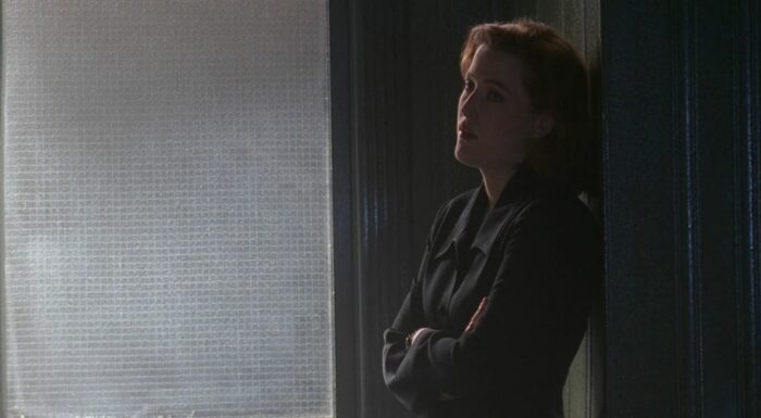 Scully leans against a wall, looking skeptical