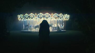 Emma, seen from behind, approaches a lit-up carousel