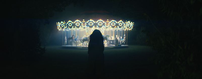 Emma, seen from behind, approaches a lit-up carousel