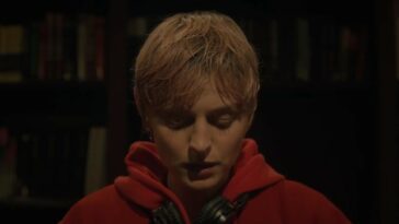 A Murder at the End of the World S1E1 - Darby with blond hair, a red sweatshirt and headphone around her neck looks down at something, wall to wall books behind her