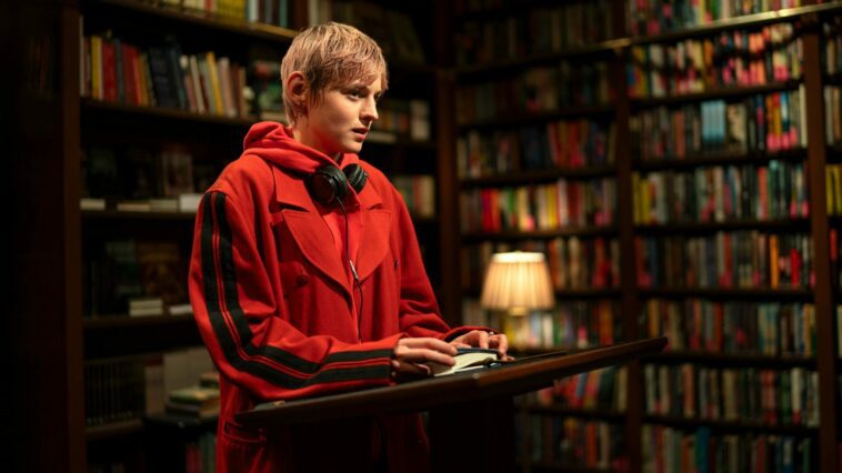 Darby stands at a podium in a bookstore in A Murder at the End of the World