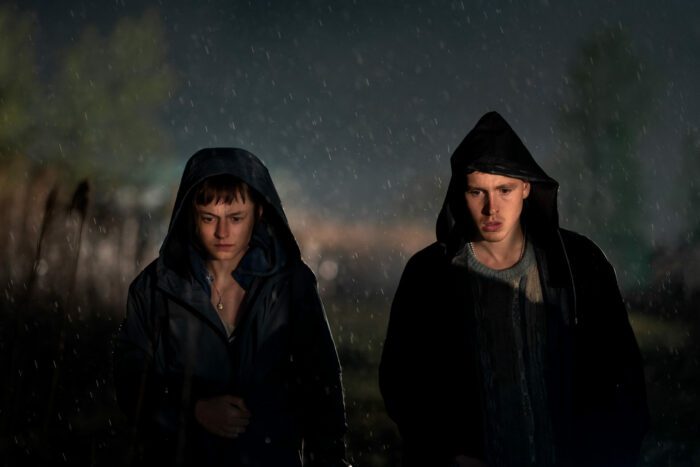 Young Darby and Bill stand in a field in raincoats with hoods